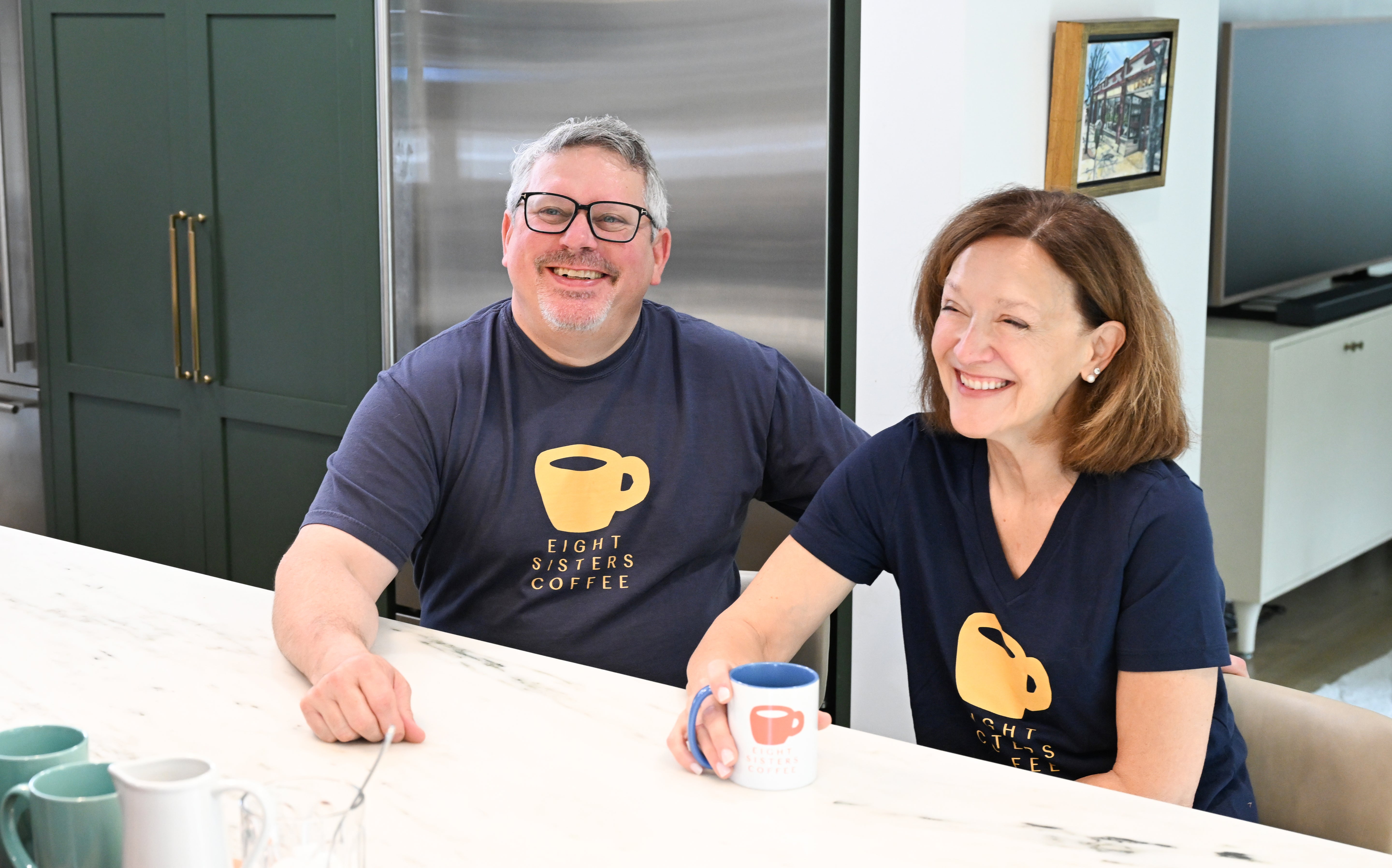 Two people sitting at a kitchen counter with coffee mugs, wearing matching navy blue t-shirts with a yellow coffee cup logo.