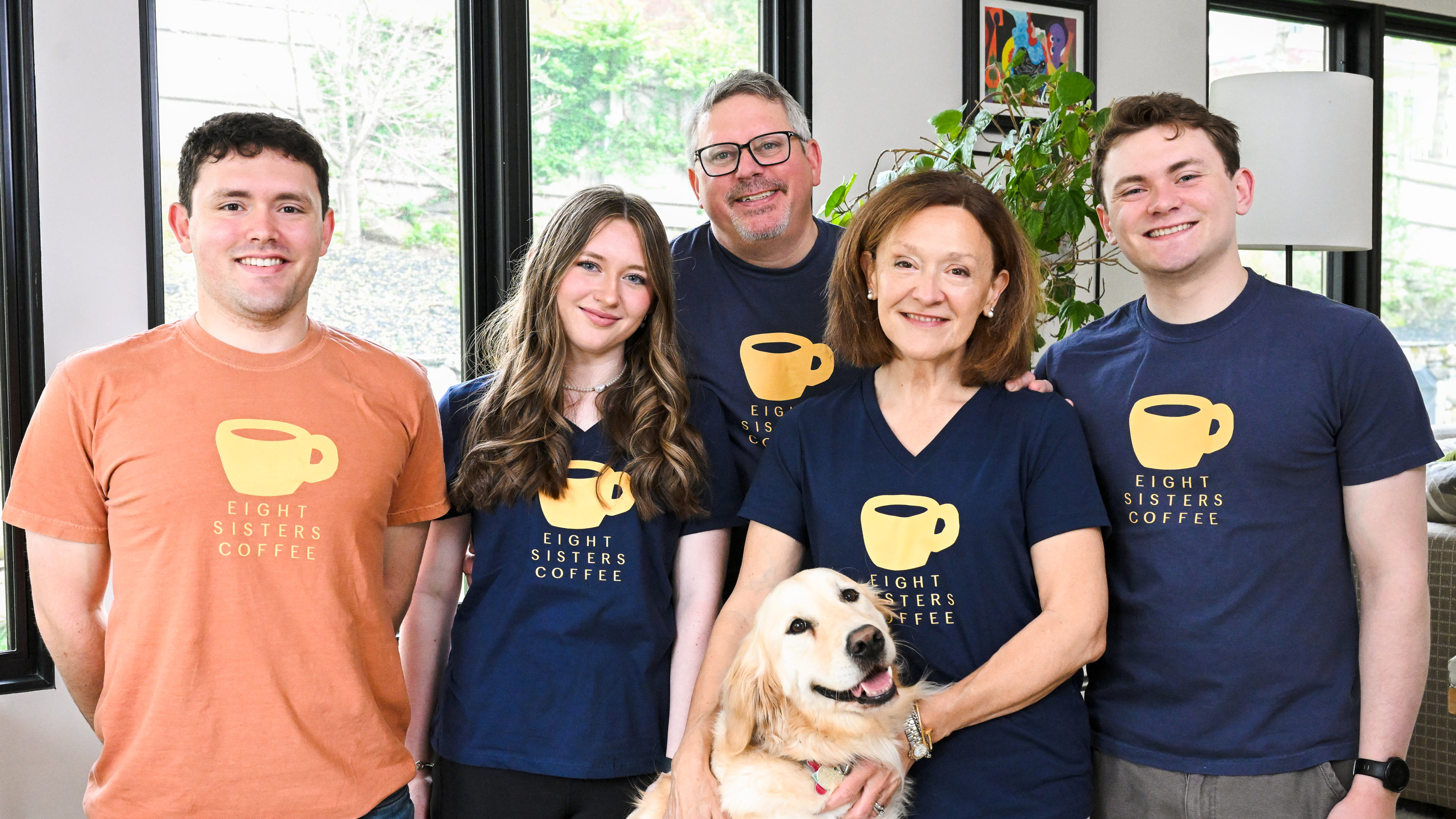 Group of people and a dog wearing 'Eight Sisters Coffee' t-shirts in an indoor setting.
