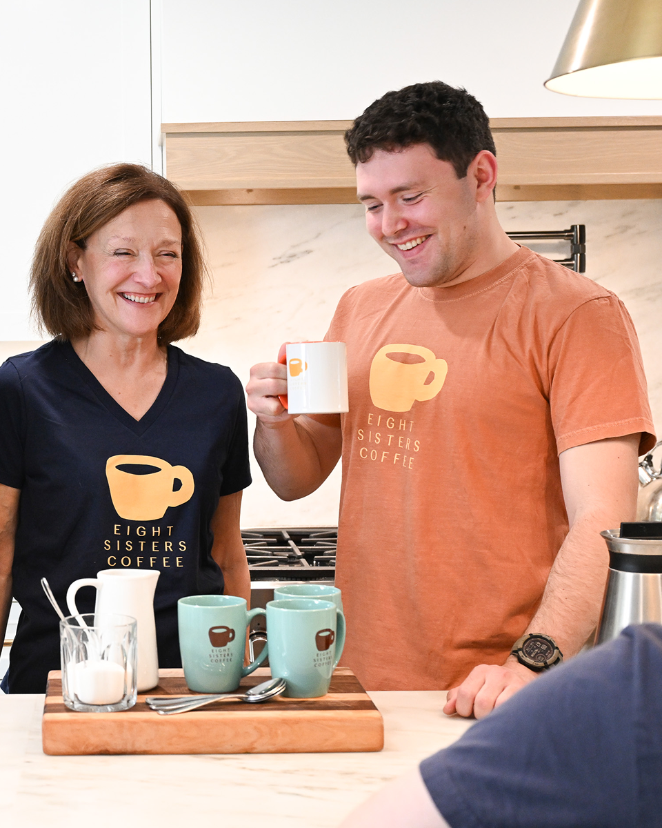 Two people in a kitchen with mugs and a cutting board, wearing branded t-shirts.