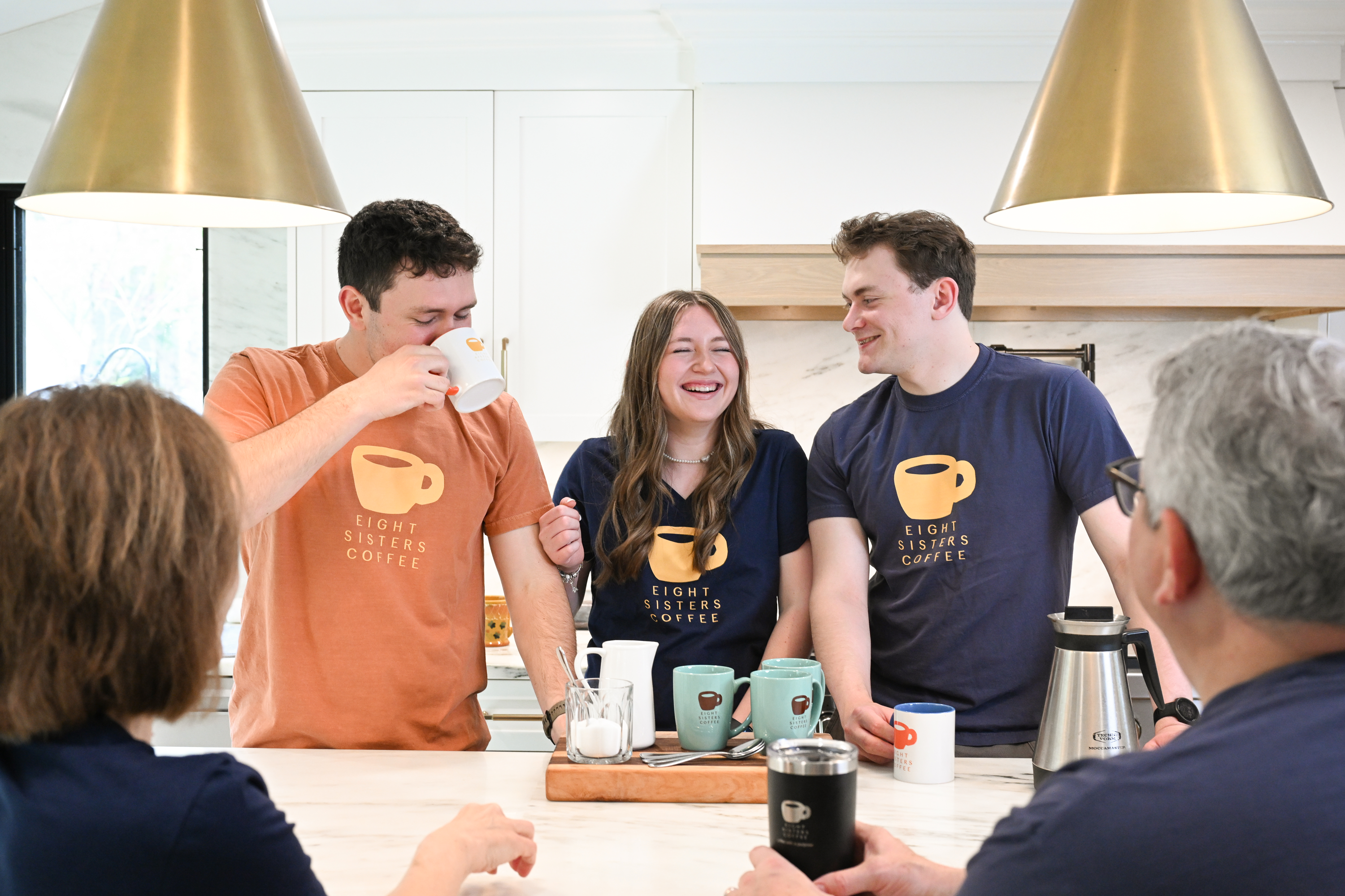 People in a kitchen setting with coffee cups and a person drinking from a mug.
